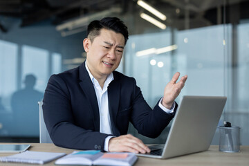 Dissatisfied young Asian male businessman sitting at desk in office and looking at laptop gesturing with hand