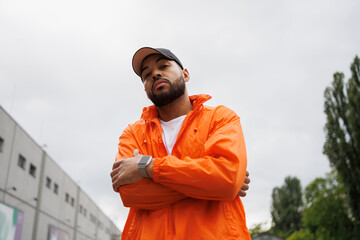 Confident african american man in windbreaker and baseball cap looking at camera on street