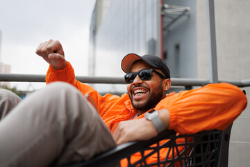 Cheerful african american man in sunglasses having fun in shopping cart on street