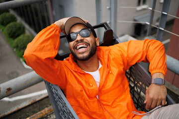 Cheerful african american man in sunglasses and windbreaker sitting in shopping cart on street