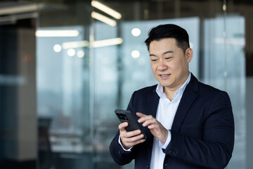 Smiling Asian young male businessman standing in office and using mobile phone