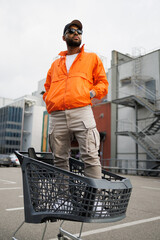 African american man in baseball cap and windbreaker jacket standing in shopping cart on street