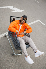 Cheerful and stylish african american man sitting in shopping cart on parking on street