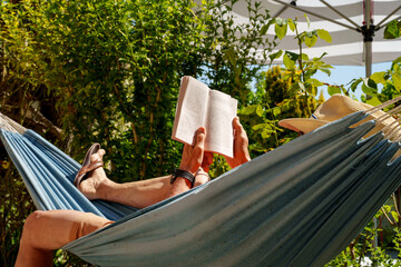 A man wearing a sun hat reads a book in a hammock. Lush garden landscape and striped parasol....