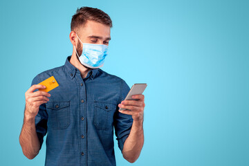Contactless shopping during covid isolation. Portrait of cool millennial guy in face mask holding smartphone and credit card over blue studio background. Young man buying goods in online store