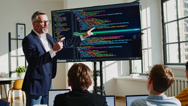 Mature software engineer pointing at computer code on a big screen, explaining it to his team of young developers sitting at a table with laptops during a meeting in a modern office