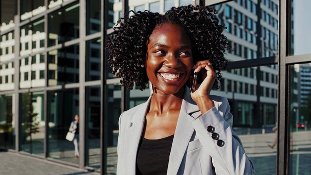 Smiling african american businesswoman enjoying a pleasant phone conversation while walking confidently near an office building in a bustling business district, radiating professionalism and style