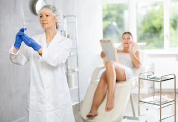 Woman nurse in medical clothes and disposable cap stands with syringe in hand and conducts...