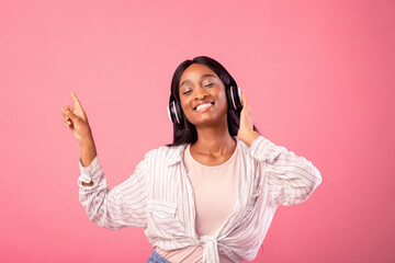 Gorgeous black lady with headset listening to music and dancing with closed eyes, pointing aside on pink studio background. Pretty African American woman moving to her favorite melody