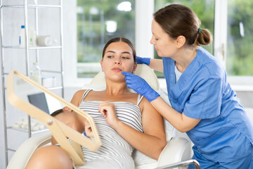 Adult female doctor cosmetologist analyzes face of young female patient in medical office