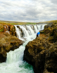 Visitors admire the stunning waterfalls cascading through Kolugljufur Canyon, surrounded by rugged cliffs and lush greenery under a dramatic sky. A perfect adventure in Iceland natural beauty.