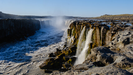 Obraz premium A powerful waterfall flows dramatically over rugged cliffs at Dettifoss, one of Icelands most breathtaking natural wonders. The sound of rushing water echoes in the serene landscape.