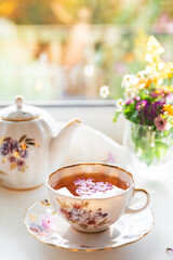 Creative postcard with porcelain cup of tea, teapot and a vibrant bouquet of garden flowers. Elegant tea set in beautiful soft sunlight. Good morning and teatime concept. Slow life visual trend.