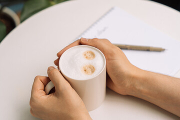 Close-up of hands holding a cup of coffee, notepad and pen in the background. High quality photo