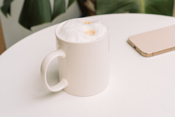 Cup of cappuccino with frothy milk on a white round table, smartphone in the background. High quality photo