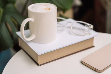Cozy home setup with cappuccino, book, eyeglasses, and smartphone on a round table. High quality photo