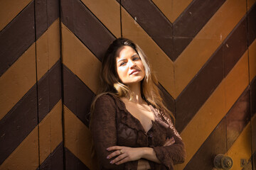 Fototapeta premium Portrait of a young, pretty woman, with brown hair, wearing a brown shirt, with her arms crossed, posing looking at the camera sweet and tender, receiving the sun's rays on her face, leaning on a door