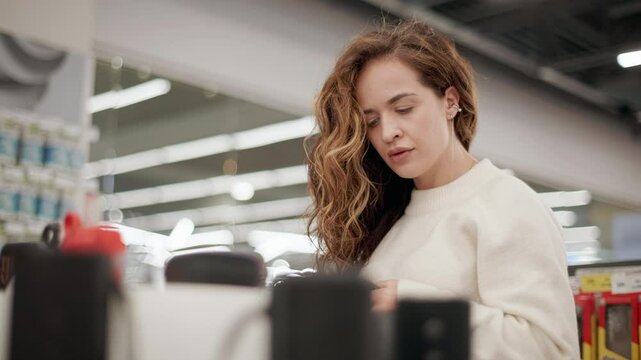 Young woman examining bluetooth speaker in electronics store, feeling happy while exploring sound technology with genuine enthusiasm and interest during shopping experience