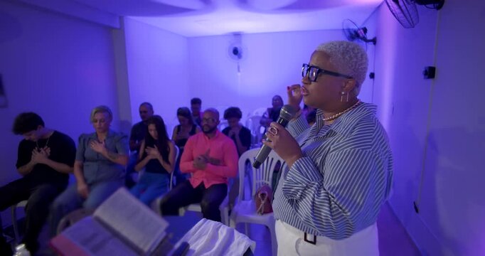 Female preacher with short blond hair passionately preaching to congregation, holding microphone in small church room with open Bible and oil bottle on table