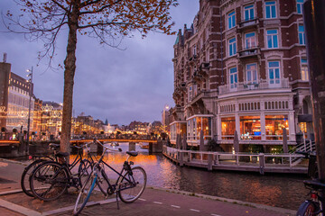 Nighttime Amsterdam. The city is illuminated with various lights. Along the canal, beautiful buildings with cafes and restaurants create a lively atmosphere. Bicycles are parked along the streets.