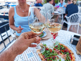 Outdoor dining scene featuring two people toasting with glasses of white wine at a seaside restaurant. In the foreground, there's a delicious pizza.