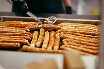 Traditional German sausages being grilled on a large tray in a professional kitchen, flipped by a cook using tongs and wearing black gloves.