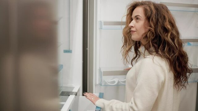 Woman carefully examining modern refrigerator in electronics store, slowly opening and closing doors and drawers while evaluating appliance features and design for potential purchase