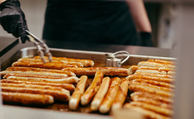 Traditional German sausages being grilled on a large tray in a professional kitchen, flipped by a cook using tongs and wearing black gloves.