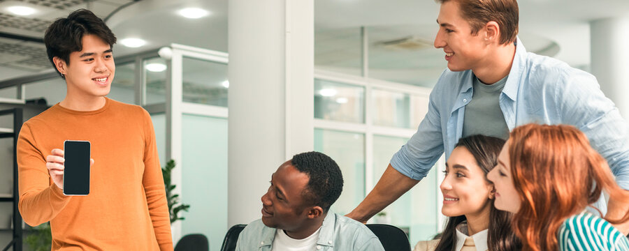 Startup Coworkers Meeting. Diverse Group Of Young Business People Gathered Together Discussing Creative Idea, Smiling Asian Man Showing Phone With Blank Screen. Brainstorming Work Process Office