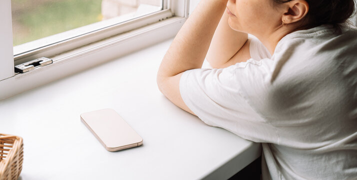 Side view of a woman daydreaming by the window with a phone lying nearby. High quality photo