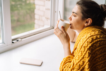 Woman sipping coffee in a knitted yellow blanket by the window, phone placed aside. High quality photo