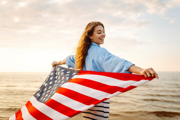 Happy woman on the beach with American flag on her shoulders. Beautiful woman enjoying sunset at...