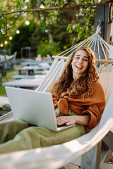 Smiling woman with laptop lying in a hammock in a sunny park. Beautiful female freelancer working...