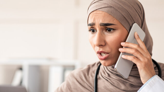 Closeup of angry female doctor giving recommendation to patient on mobile phone, young woman in hijab physician looking at laptop screen and yelling, having difficult situation at clinic, copy space