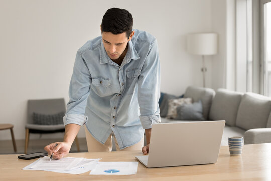 Remote work and data analysis. Busy gen z male financial analyst accountant freelancer stand by home office desk hold pen focused on reviewing paper documents prepare statistic report using notebook