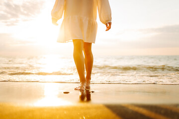 Legs of a young woman walking along the seashore at sunset. Beautiful woman in a dress on the...