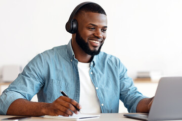 Online Education. Smiling Black Guy In Headphones Study With Laptop And Taking Notes, Cheerful...