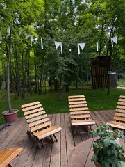 Obraz premium A charming outdoor setting featuring rustic wooden lounge chairs and a round table with a potted plant on a wooden deck, all surrounded by lush green trees and festive string lights overhead.