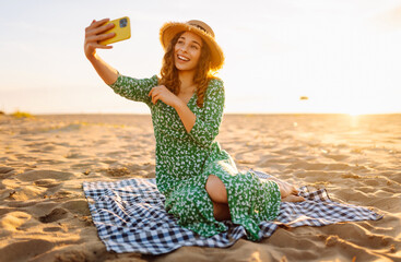 Young woman taking a selfie with a phone in her hands on the sunset beach. Cheerful woman in a...