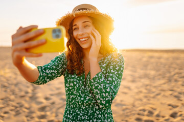 Young woman taking a selfie with a phone in her hands on the sunset beach. Cheerful woman in a bright dress and hat enjoying the seascape and chatting on her phone. Blogging and leisure concept.