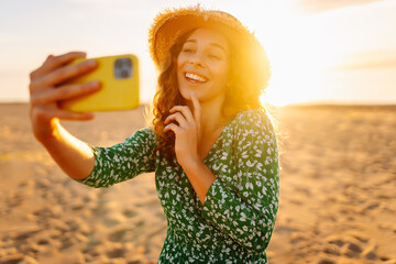 Young woman taking a selfie with a phone in her hands on the sunset beach. Cheerful woman in a...