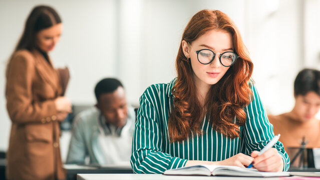 Knowledge Concept. Portrait of red-haired female student in eyeglasses sitting at desk in classroom at university, writing in notebook, taking notes, exam or test. Return to college and high school - Powered by Adobe