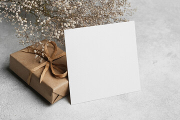 Square blank card standing on a textured surface next to a kraft paper gift box with a ribbon and...
