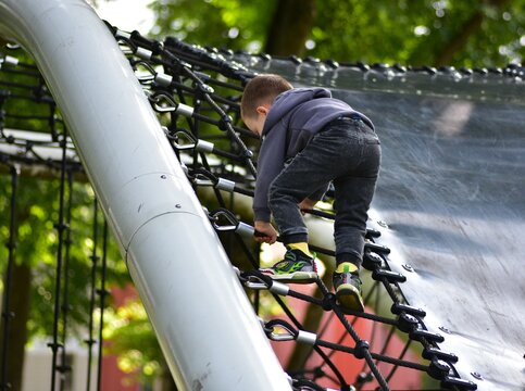 Little toddler boy in a hoodie climbs rope net at outdoor playground. Concentrated toddler enjoying adventure and physical activity in park. Child development and motor skills in action.