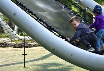 Little toddler boy in a hoodie and a girl climbs rope net at outdoor playground. Concentrated toddlers enjoying adventure and physical activity in park. Children development and motor skills.