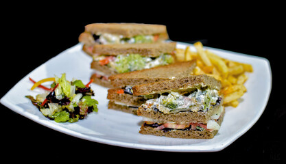 A delicious looking Brown Bread Club Sandwich layered with veggies like tomatoes, lettuce, broccoli, carrots etc with mayonnaise. Served alongside sides of French Fries and a fresh Garden Salad