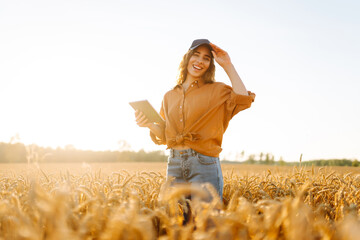 Young female farmer with digital tablet working in golden wheat field at sunset. Agronomist woman...