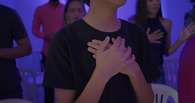 Teenage boy singing with eyes closed and hands on chest during evangelical worship service in church filled with diverse people in spiritual prayer