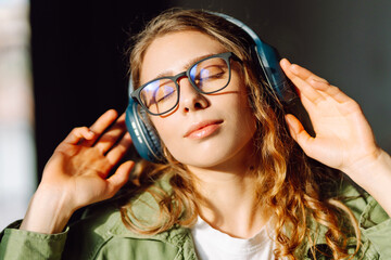 Young woman in glasses listens to music on wireless headphones and enjoys the sun in a cozy...