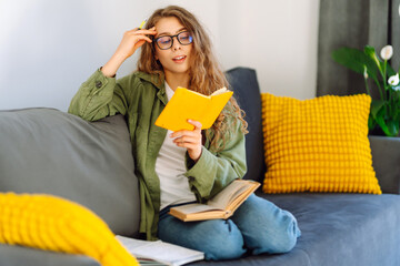 Portrait of smiling woman with yellow notebook in hands sitting on sofa in cozy room. Young female...